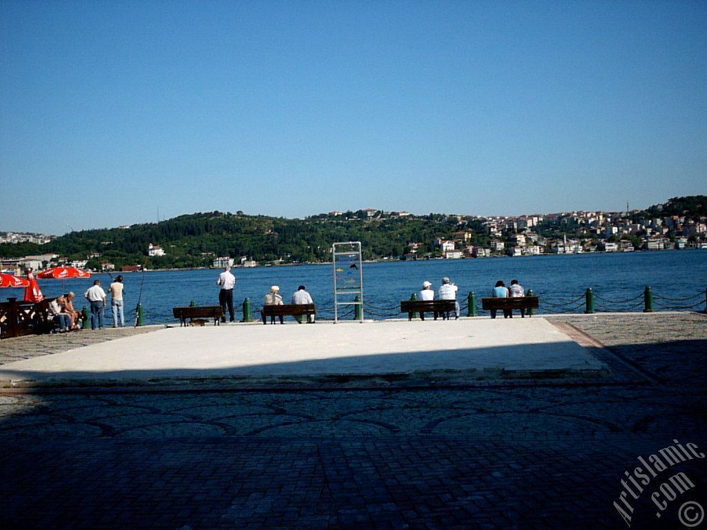 stanbul Ortaky sahilinde bir parkta oturanlar, balk tutanlar ve ufukta skdar-Beylerbeyi srtlar.
